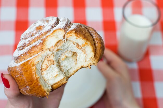 Gebissene Zimtschnecke im Frauenhandglas des karierten Tischdeckenhintergrunds der Milchweißplatte