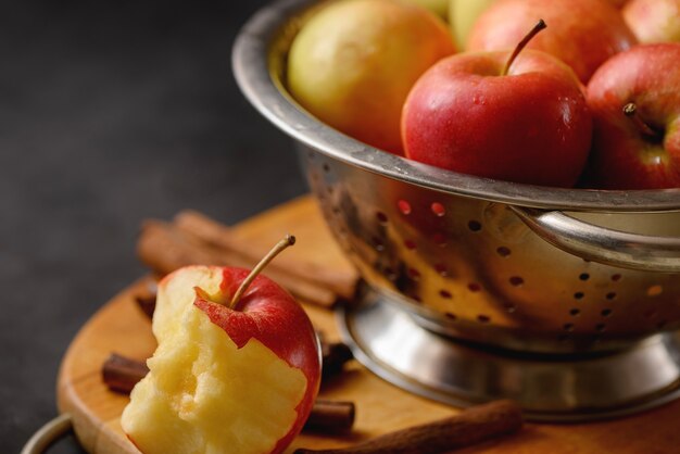 Gebissener Apfel auf Schneidebrett mit verstreuten Zimtstangen auf Metallschale voller roter reifer Äpfel.