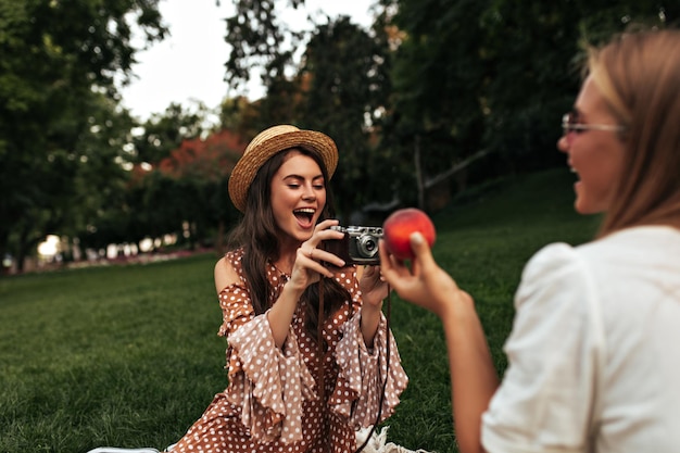 Gebräunte junge brünette und blonde Frauen in stylischen Sommerkleidern und lächelnden Bootsfahrern machen ein Picknick und trinken draußen süßes Sodawasser