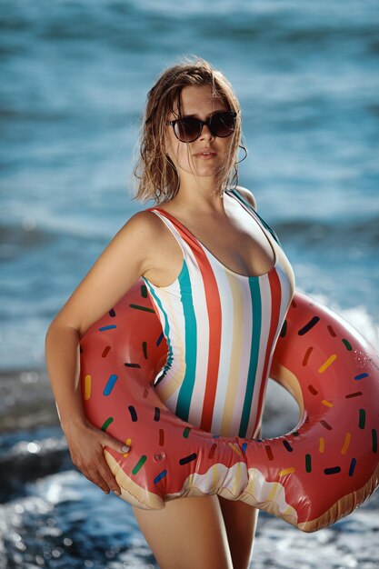 Gebräunte junge Frau in Badeanzug und Sonnenbrille, die an einem Strand mit einem aufblasbaren Donut-Ring steht