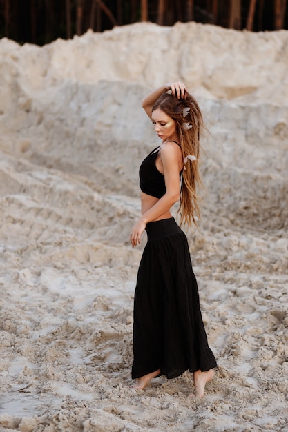 Gebräuntes Mädchen in einem schwarzen Rock und T-Shirt mit Dreadlocks im Sand bei Sonnenuntergang im Sommer