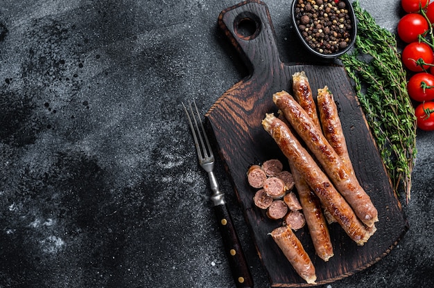 Gebratene Fleischwurst auf einem Holzbrett mit Kräutern und Tomaten. Schwarzer Hintergrund. Ansicht von oben. Platz kopieren.