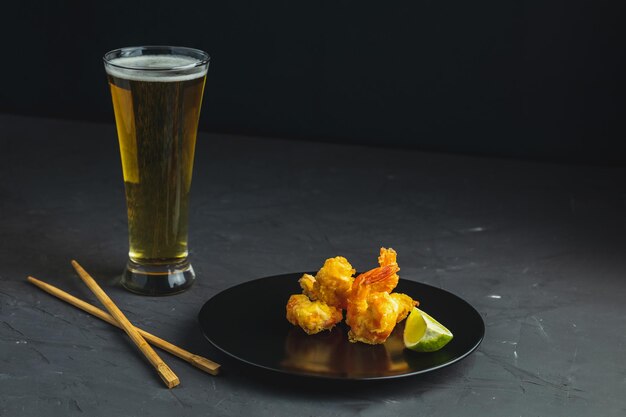 Foto gebratene garnelen-tempura mit limette auf schwarzem teller und ein glas helles bier auf dunklem betonhintergrund. kopierraum. meeresfrüchte-tempura-gericht, serviert im japanischen oder ostasiatischen stil mit stäbchen