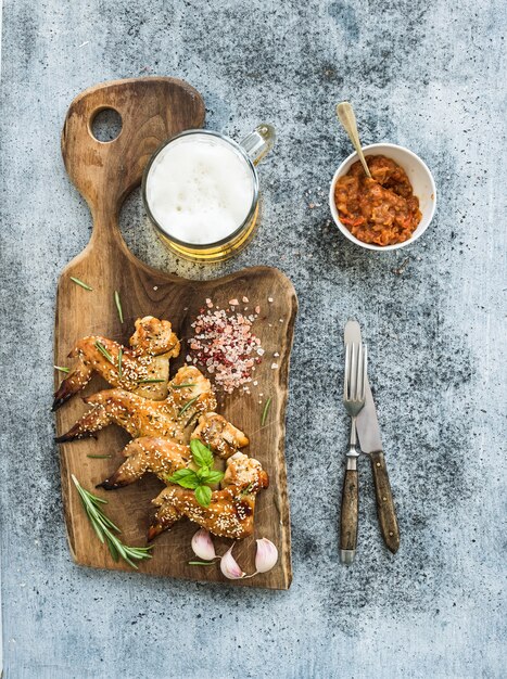 Gebratene Hühnerflügel auf rustikalem Umhüllungsbrett, würziger Tomatensauce, Kräutern und Becher hellem Bier über schwarzem hölzernem Hintergrund