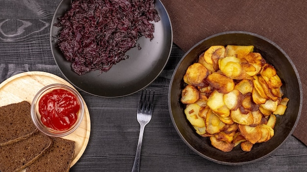 Gebratene Kartoffelchips in einem Topf mit dunklem Roggenbrot auf einer Holzplatte und hausgemachtem gedünstetem Kohl, auf schwarzem rustikalem Hintergrund. Ansicht von oben