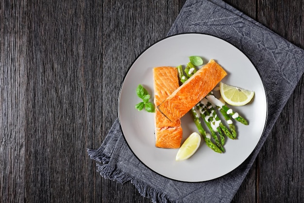 Gebratene Lachsfilets mit Zitronenspalten und gegrilltem Spargel, bestreut mit Sauce Hollandaise und auf einem Teller auf einem dunklen Holztisch lagen flach auf freiem Platz