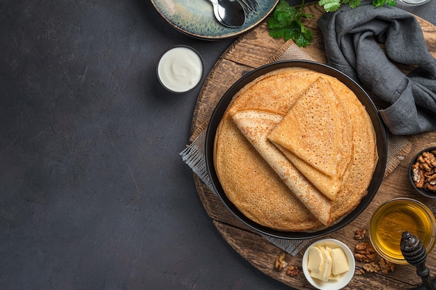 Gebratene Pfannkuchen auf dunklem Hintergrund mit Honig, Sauerrahm und Nüssen. Pfannkuchentag. Ansicht von oben, Kopienraum.