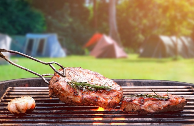 Gebratene, reife Rindersteaks auf flammendem Holzkohlegrill mit verschwommenem Hintergrund des Campingplatzes