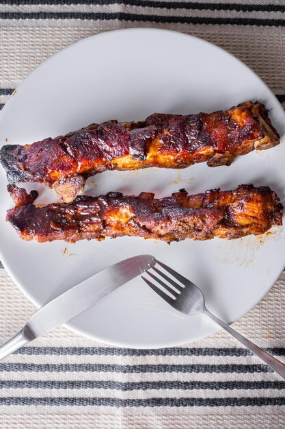 Gebratene Schweinerippchen mit Barbecue-Sauce auf einem weißen Porzellanteller mit einem Graphit und einem Messer. Gestreifte weiße Tischdecke