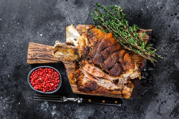 Gebratene Schweinshaxe Eisbein auf einem Holzbrett mit Kräutern. Schwarzer Hintergrund. Ansicht von oben.