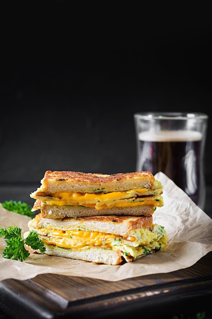 Gebratener Sandwichkäse und Rührei auf Papier mit Kaffeeglastasse auf Holzbrett. Dunkler Hintergrund.