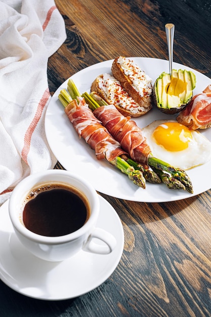 Gebratener Spargel, eingewickelt in Jamon Spiegeleier Bruschetta mit Weichkäse und Avocado auf einem weißen Teller und einer Tasse frisch gebrühtem Kaffee