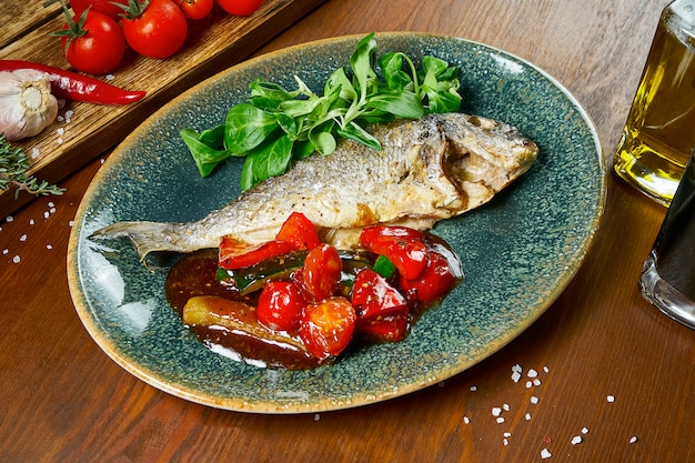 Gebratenes Fischdorado mit Gewürzen und Olivenöl, garniert mit Spinat, Tomaten und Gurken in süß-saurer Sauce auf einem blauen Teller auf einem Holztisch. Restaurant servieren. Nahansicht