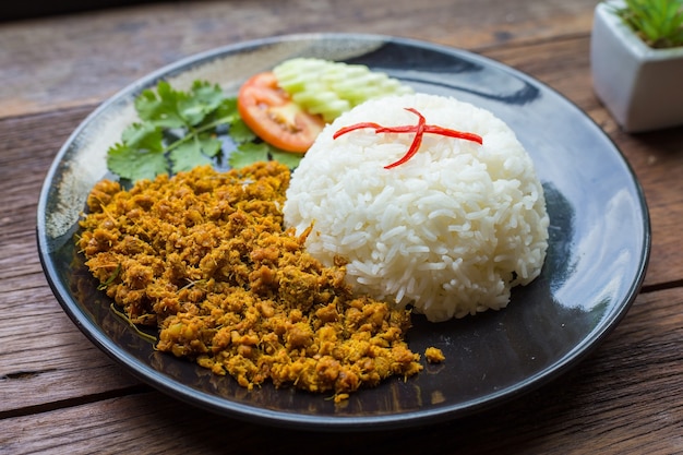 Gebratenes Schweinefleisch mit gelber Currypaste über Reis ist auf einem schwarzen Teller und einem braunen Holzhintergrund