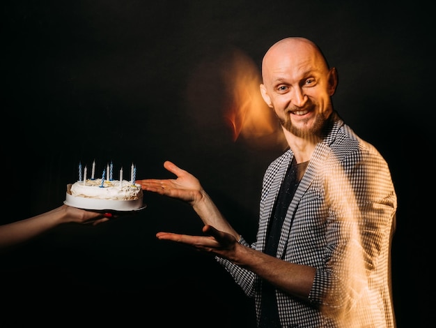 Geburtstagsmann Festlicher Kuchen Feiertagsgruß Partyfeier Fröhlicher, ausdrucksstarker, stylischer Typ, der erstaunt ist, wie er Gebäck mit Kerzenlicht bekommt, isolierte schwarze Langzeitbelichtung, defokussierter Formkopierraum