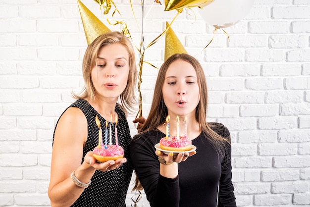 Geburtstagsparty. zwei lächelnde junge frauen oder schwestern in geburtstagshüten, die geburtstag feiern, die donuts mit kerzen über weißem backsteinmauerhintergrund halten