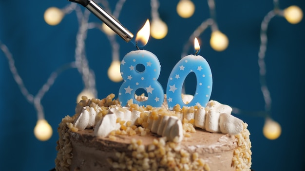 Geburtstagstorte mit 80er Kerze auf blauem Hintergrund mit Feuerzeug in Brand gesetzt. Nahaufnahme