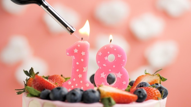 Geburtstagstorte Nummer 18 rosa Kerze auf schönem Kuchen mit Beeren und Feuerzeug mit Feuer vor dem Hintergrund von weißen Wolken und rosa Himmel Detailansicht