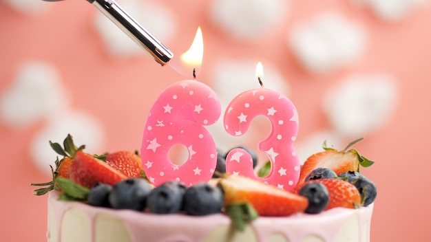 Geburtstagstorte Nummer 63 rosa Kerze auf schönem Kuchen mit Beeren und Feuerzeug mit Feuer vor dem Hintergrund von weißen Wolken und rosa Himmel Detailansicht