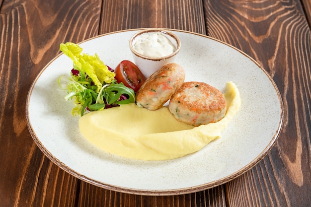 Gedämpfte Fischkoteletts mit Kartoffelpüree, Gemüse und weißer Sauce auf Holztisch