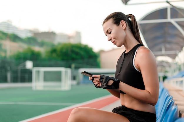 Geeignete sportliche Frau benutzt Telefon am Stadion morgens während des Eignungstrainings