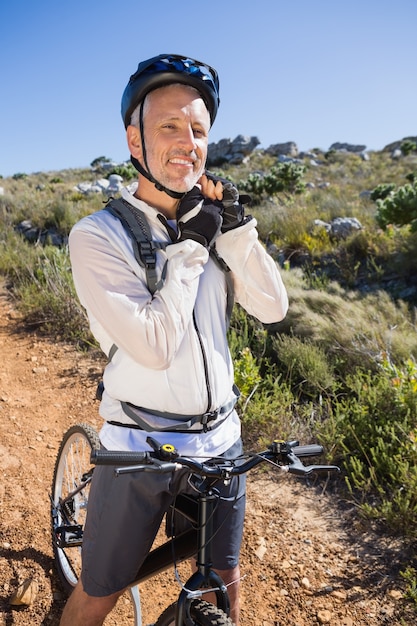 Geeigneter Radfahrer, der Sturzhelmbügel auf dem Landgelände lächelt an der Kamera justiert