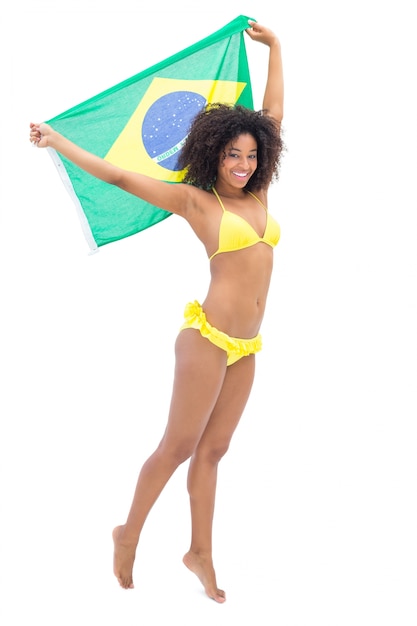 Geeignetes Mädchen im gelben Bikini, der Brasilien-Flagge lächelt an der Kamera hält