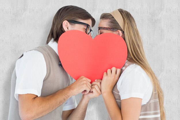 Geek hipsters besándose detrás de la tarjeta del corazón contra el fondo blanco.