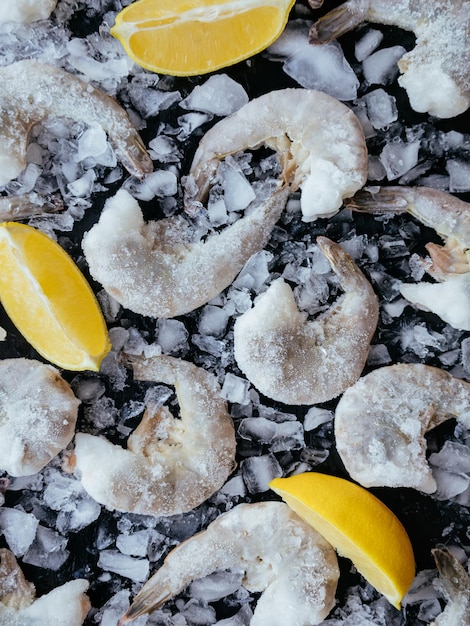 Gefrorene Ozean-Jumbo-Garnelen mit Eis und Zitrone auf schwarzem Hintergrund zum Kochen. Pazifische weiße Garnelen oder Riesengarnelen ohne Kopf für die Essenszubereitung. Meeresfrüchte-Hintergrund für die Präsentation Ihrer Produkte