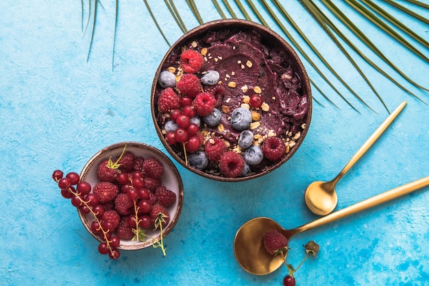 Gefrorener Acai-Smoothie in Kokosnussschale mit Himbeeren, Banane, Blaubeeren, Obst und Müsli auf konkretem Hintergrund. Frühstück, gesunde Mahlzeit für Sommerstimmung, Draufsicht, Platz für Text