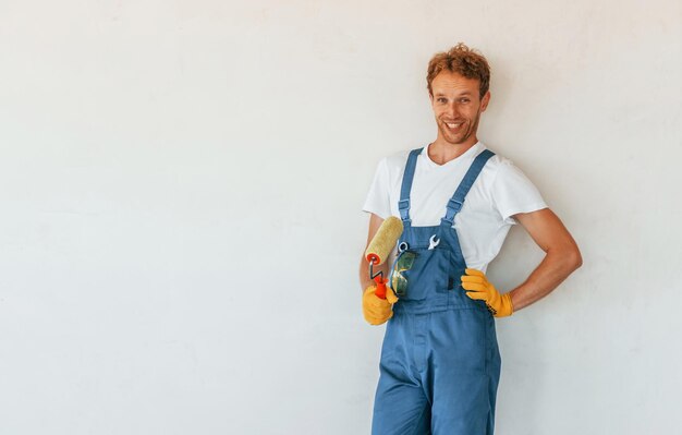 Gegen fertige weiße Wände stehend Junger Mann, der tagsüber in Uniform am Bau arbeitet