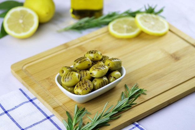 Gegrillte grüne Oliven mit Rosmarin, Olivenöl und Zitrone in einem Teller auf einem Holzschneidebrett auf dem Tisch