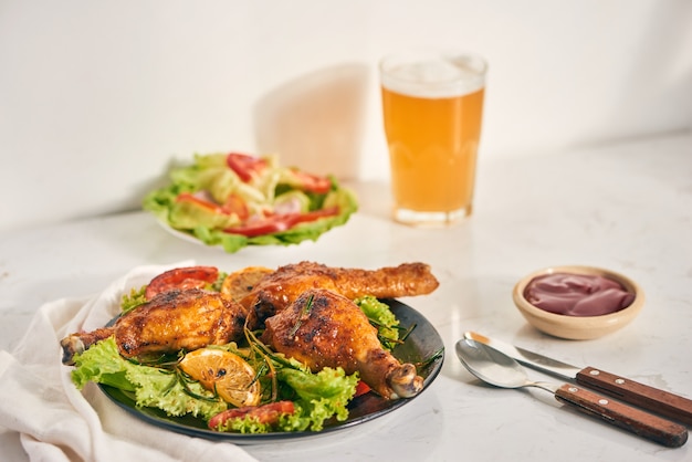 Gegrillte Hähnchenschenkel gebraten auf dem Grill auf dunklem Teller mit Tomatensauce in einer Schüssel und Salatblättern, Glaskrug Bier