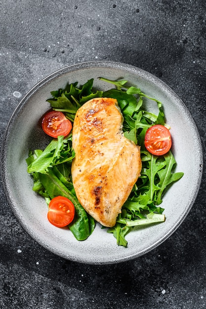 Gegrillte Hühnerbrust. Hähnchenfilet und frischer Gemüsesalat mit Tomaten und Rucola-Blättern. Schwarzer Hintergrund. Draufsicht