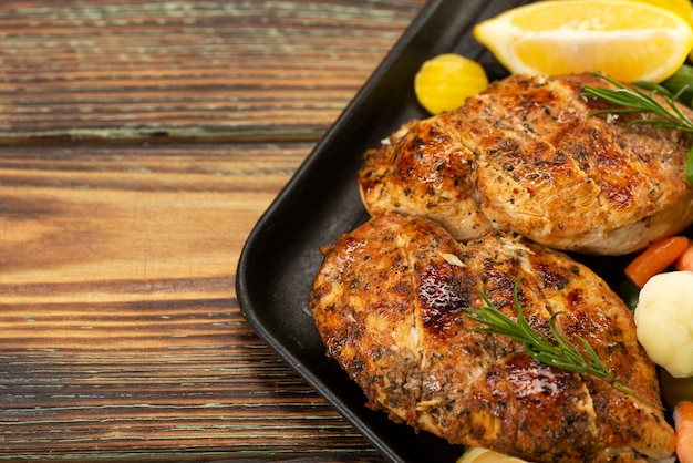 Gegrillte Hühnerbrust in einer Pfanne mit Gemüse und Kräutern und Zitronenscheiben auf einem Holzbrett