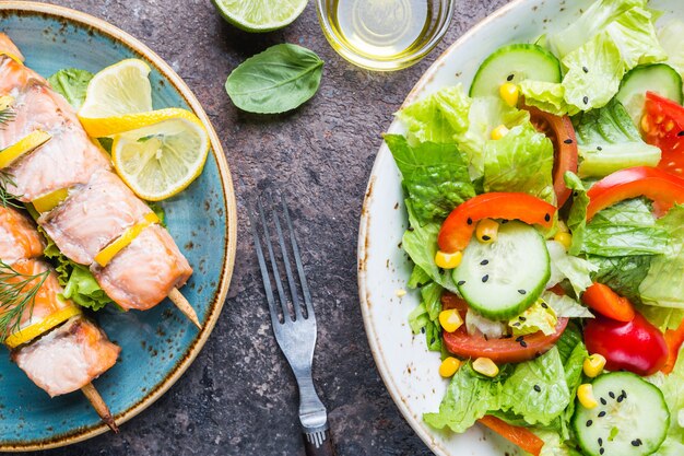 Gegrillte Lachsspieße und gesunder Gemüsesalat aus frischen Tomaten, Gurken, Salat und Mais auf Teller, Draufsicht. Diät-Menü.