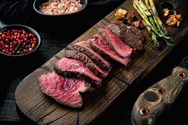 Gegrillte Rinderfiletsteaks, mit Zwiebeln und Spargel, auf Holzbrett, mit Fleischmesser und Gabel, auf schwarzem Holztischhintergrund