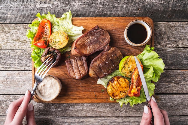 Gegrillte Rinderzunge serviert mit Gemüse und Sauce auf einem Holzbrett. Ansicht von oben