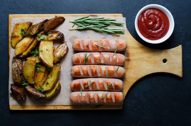 Gegrillte Würste auf einem hölzernen Schneidebrett. Bratkartoffeln, Rosmarin, Tomatenketchup. Ungesunde Diät. Dunkler Hintergrund