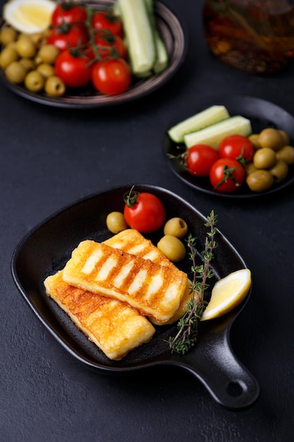 Gegrillter Halloumi-Käse in einer schwarzen Pfanne mit Oliven, Tomaten, Gurken, Thymian und Zitrone Traditioneller Käse des östlichen Mittelmeerraums. Schwarzer Hintergrund, selektiver Fokus, Nahaufnahme