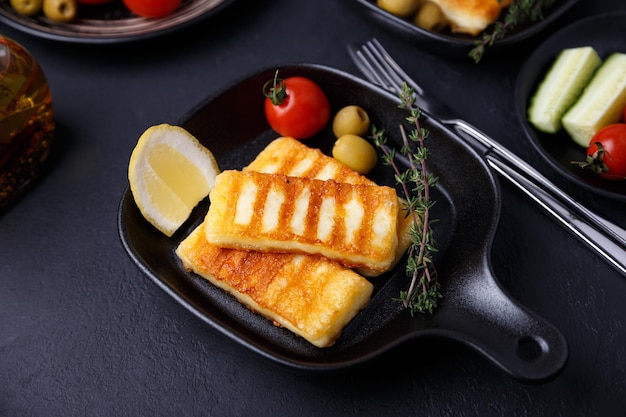 Gegrillter Halloumi-Käse in einer schwarzen Pfanne mit Oliven, Tomaten, Gurken, Thymian und Zitrone Traditioneller Käse des östlichen Mittelmeerraums. Schwarzer Hintergrund, selektiver Fokus, Nahaufnahme