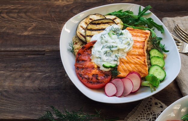 Gegrillter Lachs, Auberginen und Tomaten mit Quinoa- und Tzatziki-Sauce auf rustikalem Holz. Gesundes Abendessen.