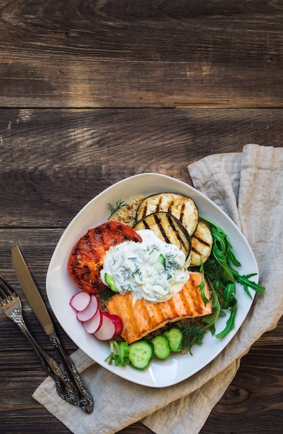 Gegrillter Lachs, Auberginen und Tomaten mit Quinoa- und Tzatziki-Sauce auf rustikalem Holzhintergrund. Gesundes Abendessen. Draufsicht. Speicherplatz kopieren.