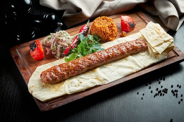 Gegrillter türkischer Adana-Kebab mit gegrilltem Gemüse, Zwiebeln und Reis auf einem Holzbrett. Dunkler Tisch.