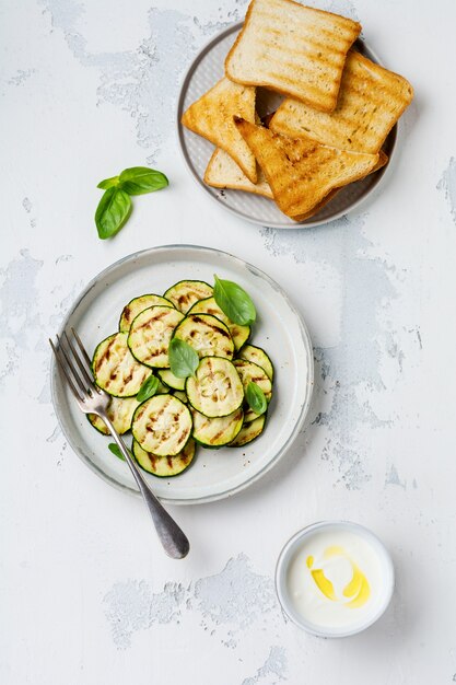 Gegrillter Zucchinisalat mit Basilikumblättern, Joghurtsauce und gebratenem Brot in einer einfachen Keramikplatte auf einer weißen Betonoberfläche