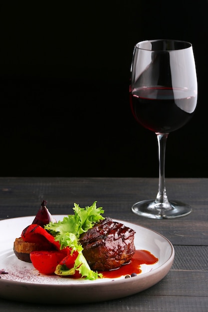 Gegrilltes Filet Mignon mit Gemüsesalat und Glas Rotwein hautnah. Teller mit Filet Mignon auf Holztisch und schwarzem Tisch