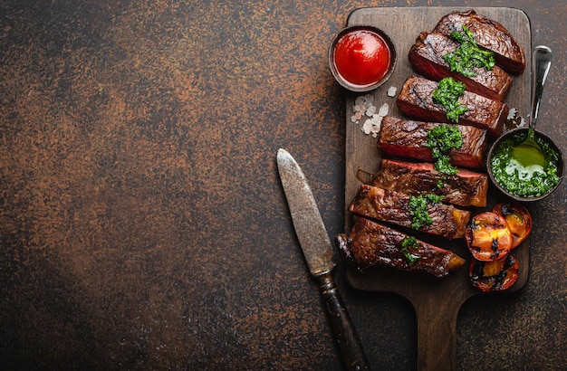 Gegrilltes/gebratenes und in Scheiben geschnittenes marmoriertes Fleischsteak mit Gabel, Tomaten, verschiedenen Saucen auf Holzbrett, Draufsicht, Nahaufnahme mit Platz für Text, rustikaler Steinhintergrund. Rindfleisch Steak-Konzept