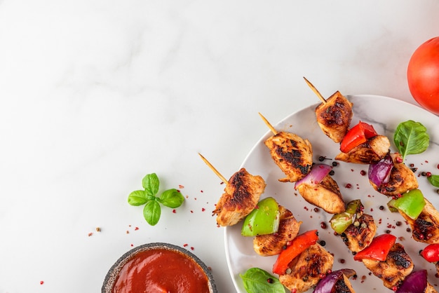 Gegrilltes Grillfleisch am Spieß mit Gemüse, Gewürzen, Basilikum und Tomatenketchup auf einem Teller über weißer Oberfläche