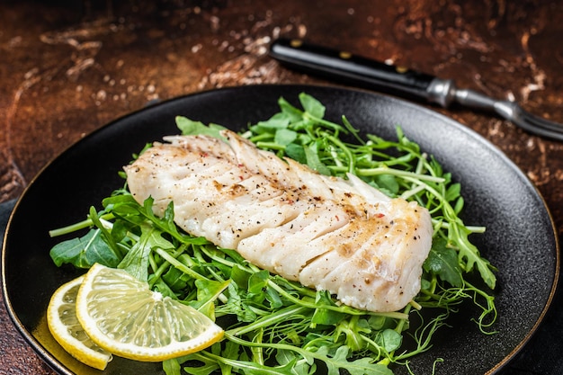 Foto gegrilltes kabeljaufilet serviert mit grünem salat in einem teller dunkler hintergrund ansicht von oben