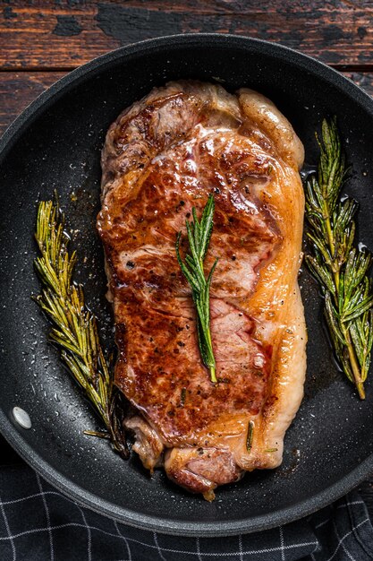 Gegrilltes New Yorker Rindfleischsteak in einer Pfanne mit Kräutern. Dunkler hölzerner Hintergrund. Ansicht von oben.